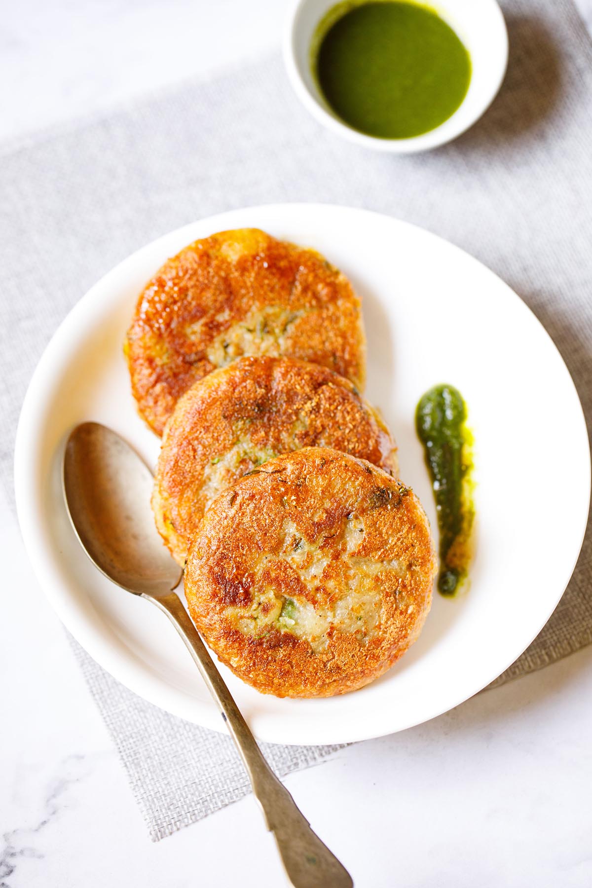 three aloo tikki kept vertically on a white plate with a spoon and a splash of cilantro dip on the plate placed on a light grey fabric.