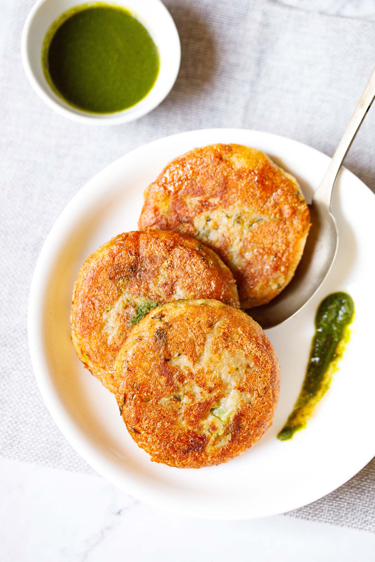 three aloo tikki kept vertically on a white plate with a spoon and a splash of cilantro dip on the plate placed on a light grey fabric.