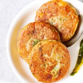 three aloo tikki kept vertically on a white plate with a spoon and a splash of coriander chutney on the plate.