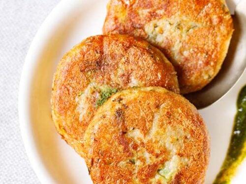 three aloo tikki kept vertically on a white plate with a spoon and a splash of coriander chutney on the plate.