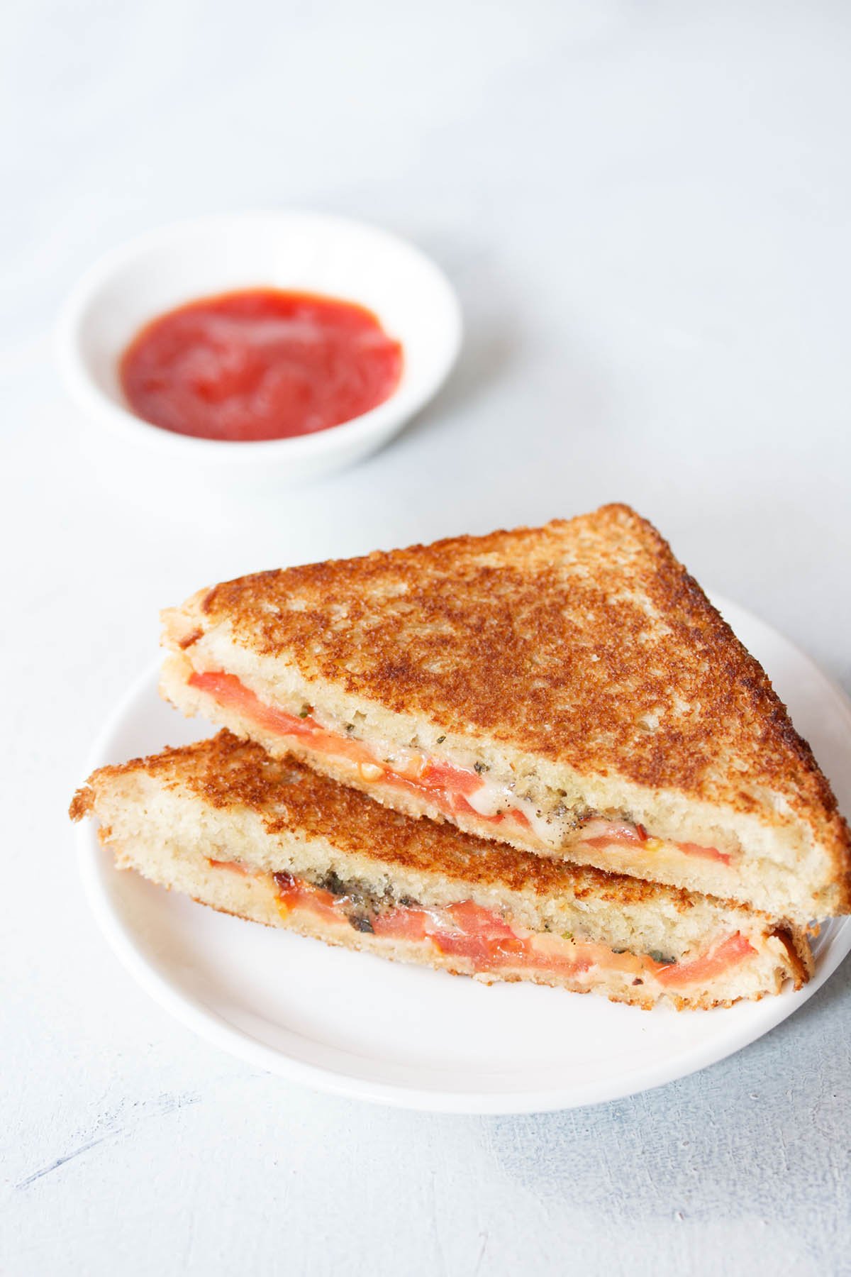 tomato sandwich served on a white plate with a small bowl of tomato sauce kept on the top side.