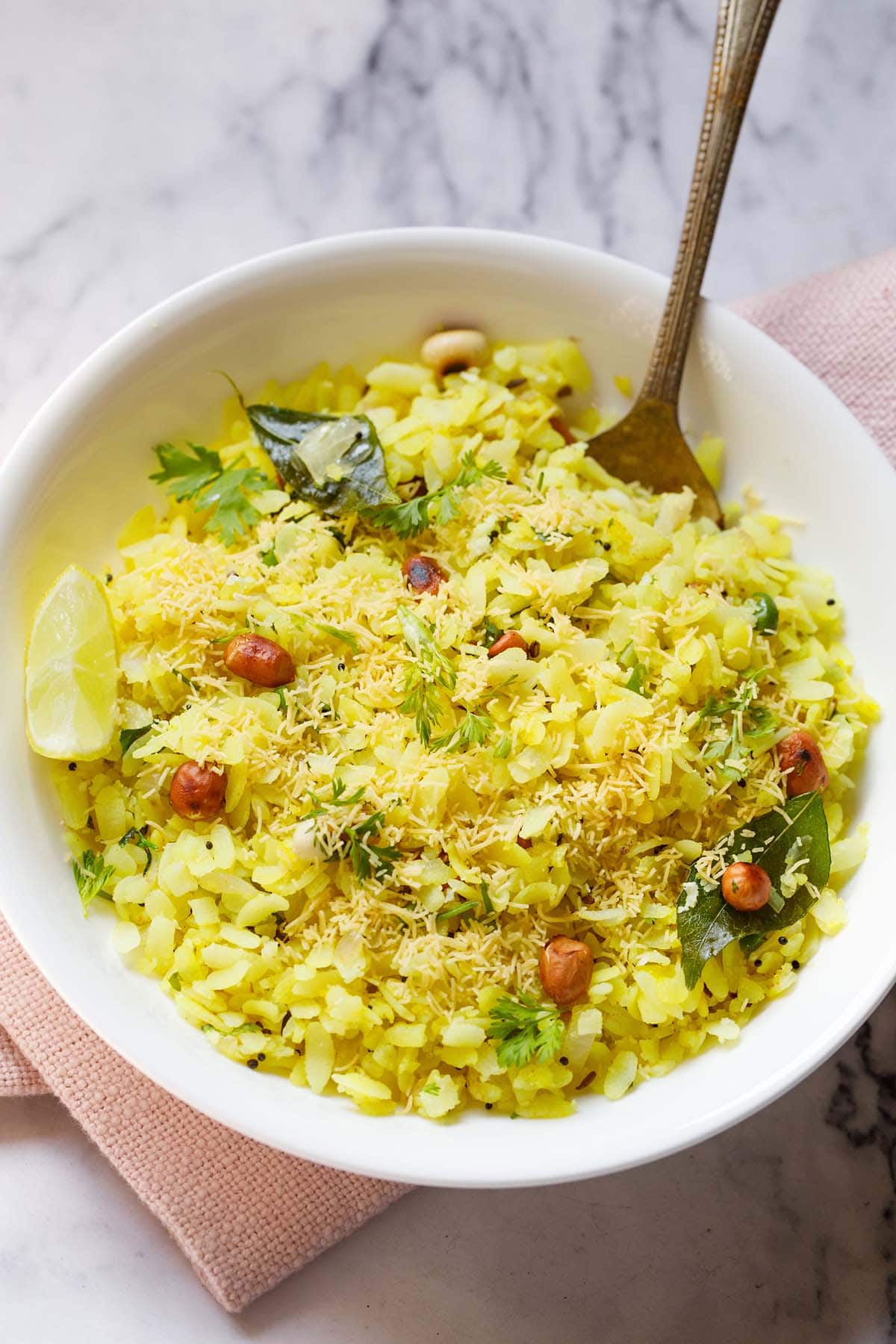 kande pohe in a white bowl with a spoon and lemon wedge inside.