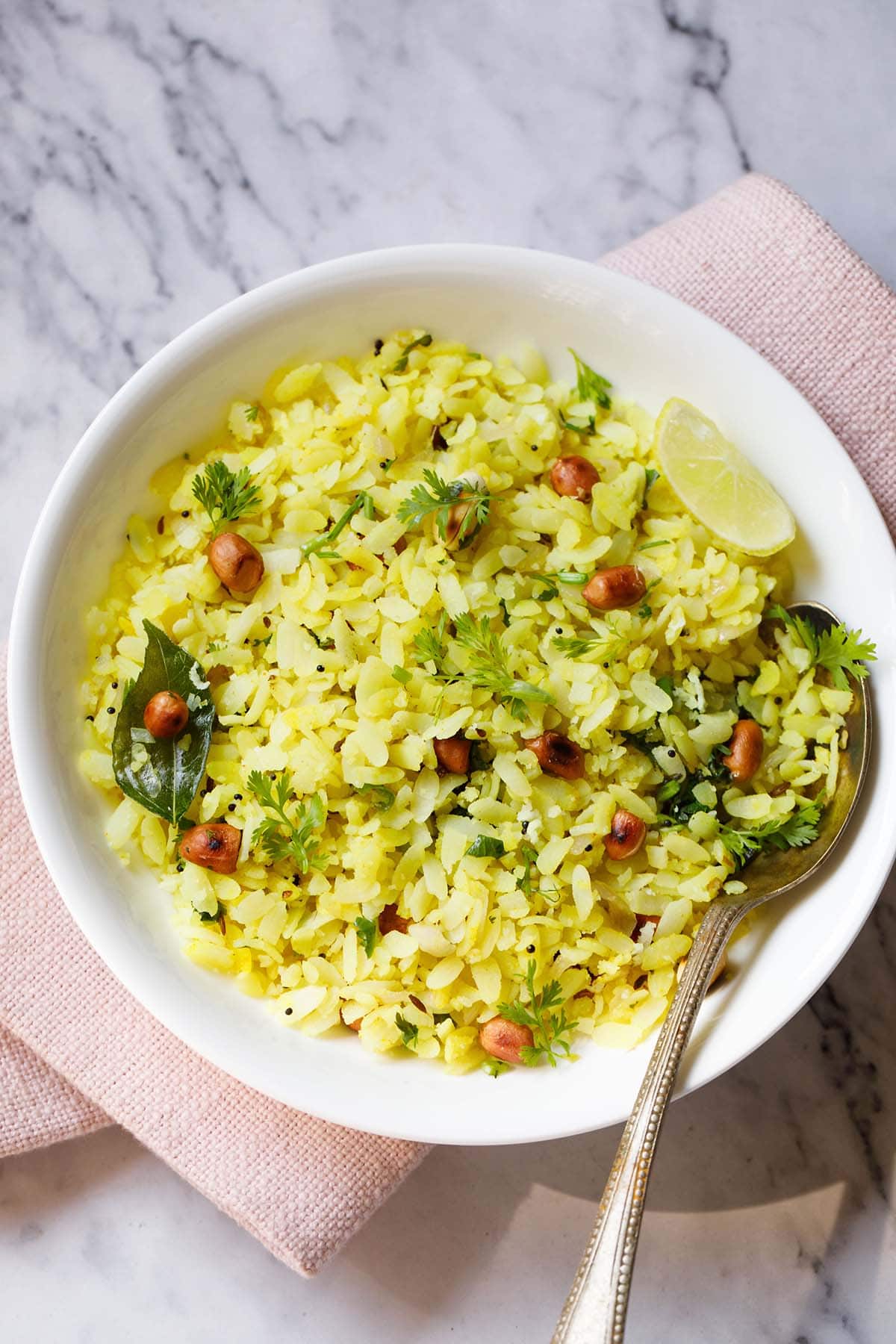 poha or pohe in a white bowl with a lemon wedge and spoon.