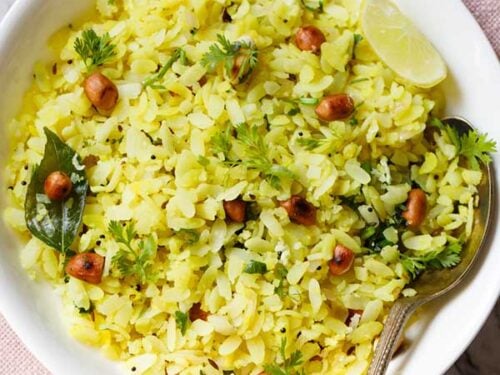 kanda poha or pohe in a white bowl with a lemon wedge and spoon.