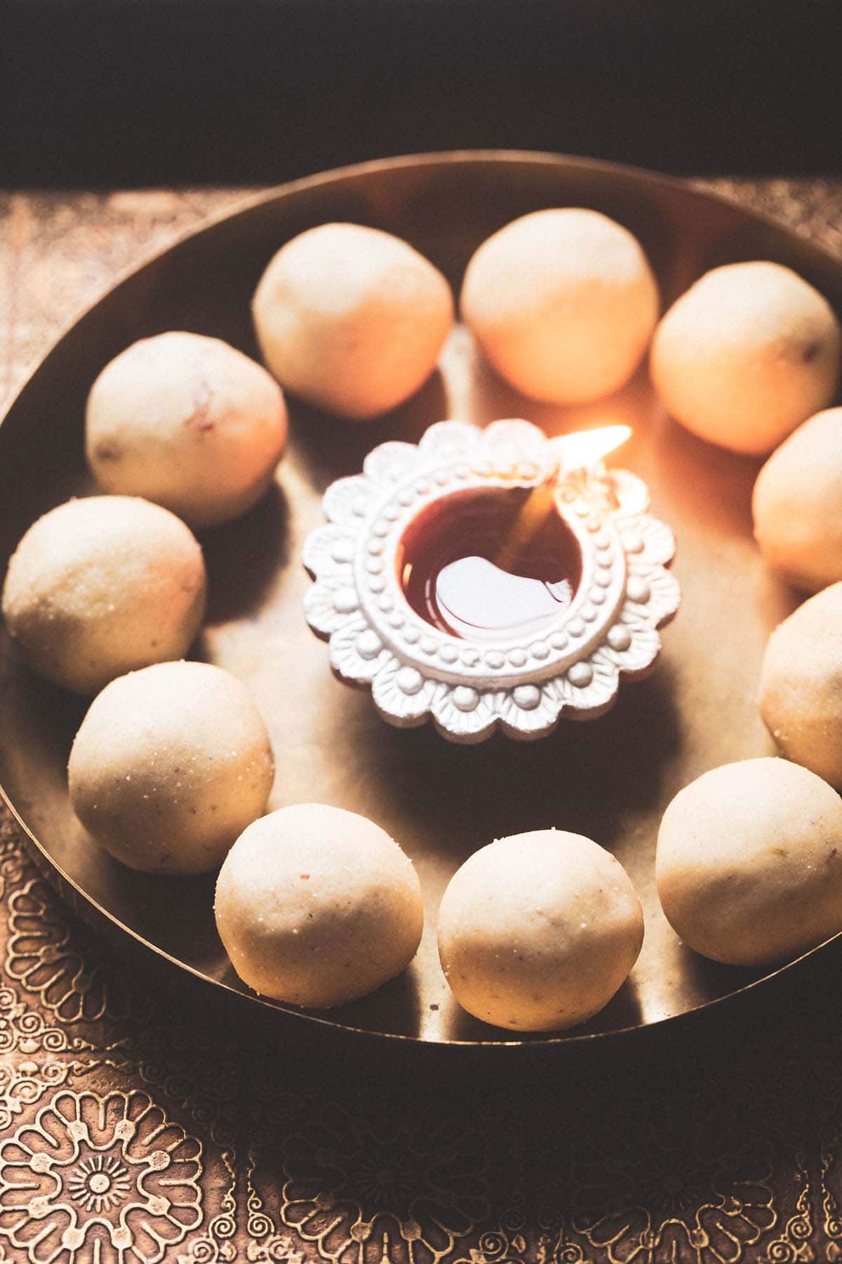 suji ke laddu in a circle with lit earthen lamp in center on a bronze plate and text layovers. 
