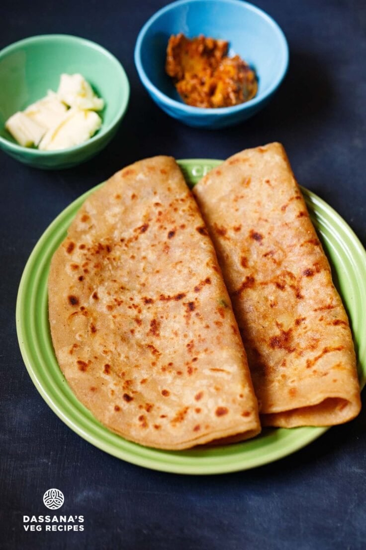 aloo paratha folded and placed on a green ceramic plate on a dark bluish board. sides of butter cubes and mango pickle served in two small ceramic bowls.