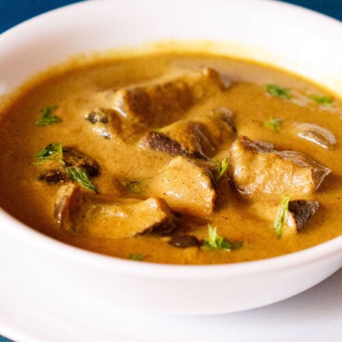 White bowl of bagara brinjal on a white plate on a blue table.