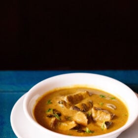 White bowl of bagara brinjal on a white plate on a blue table.