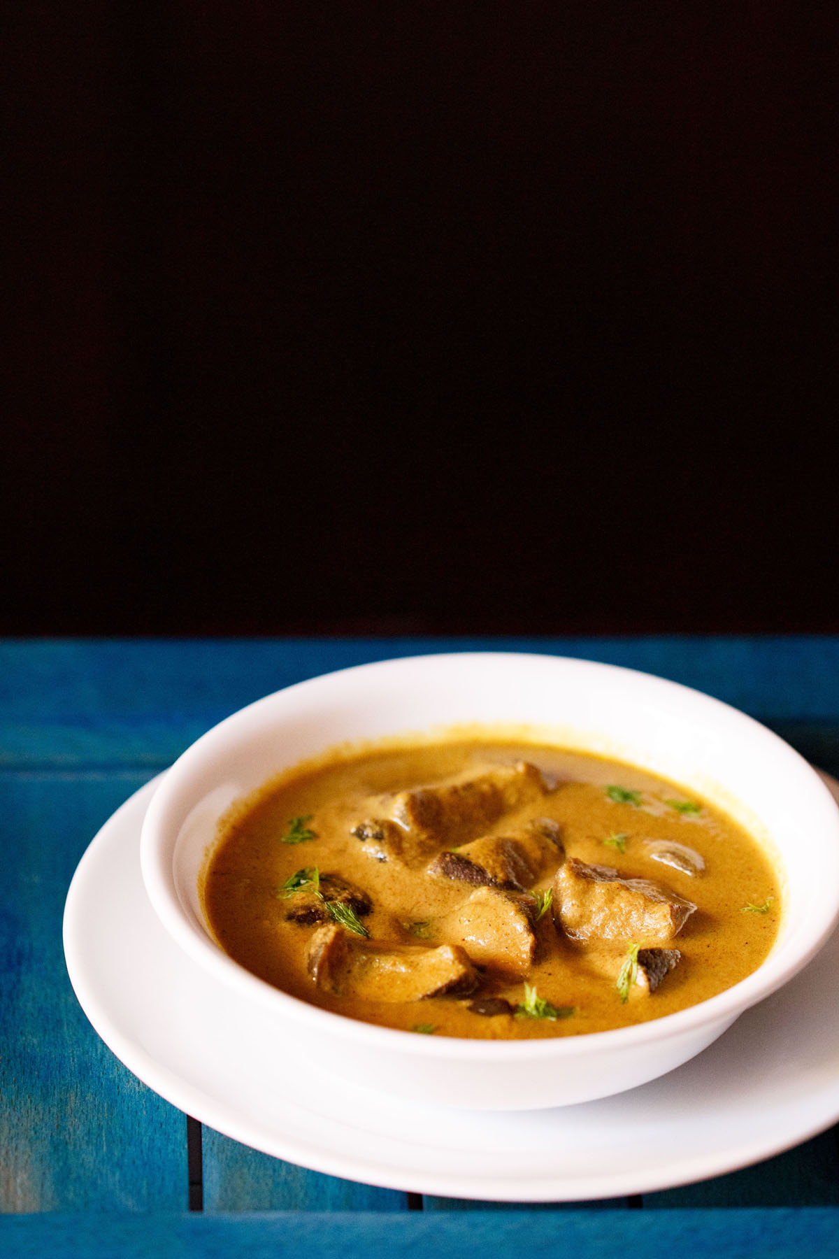 White bowl of bagara brinjal on a white plate on a blue table.