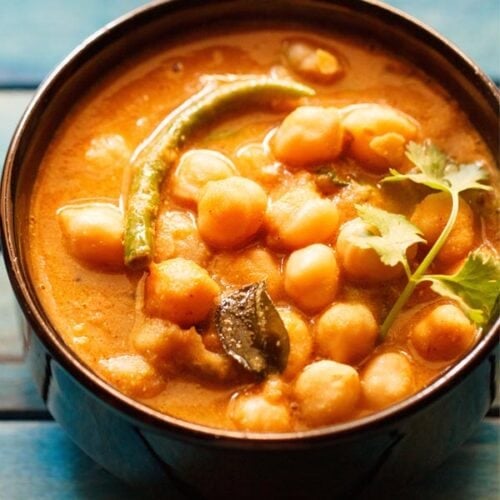 chickpea curry with a coriander sprig on top, in a black bowl on a blue wooden tray.