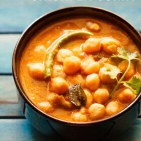 chickpea curry with a coriander sprig on top, in a black bowl on a blue wooden tray.