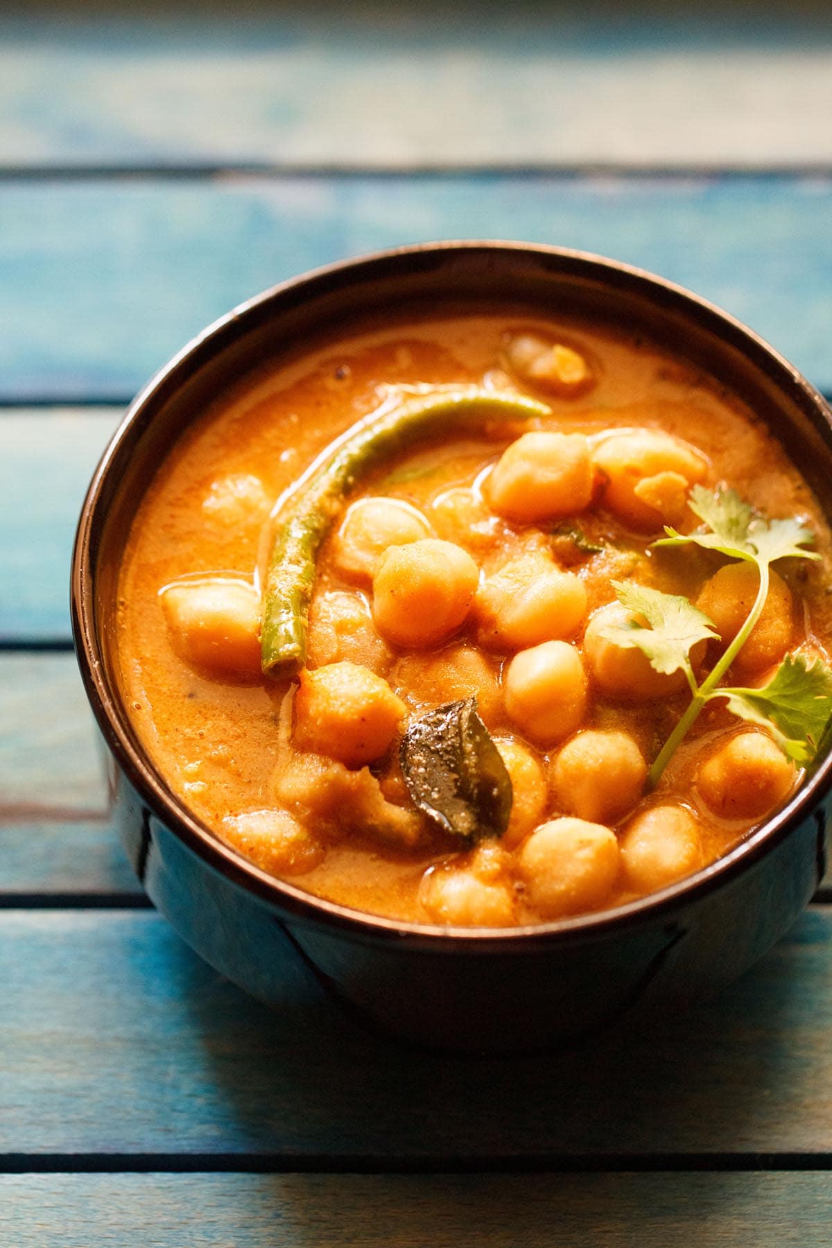 chickpea curry with a coriander sprig on top, in a black bowl on a blue wooden tray.