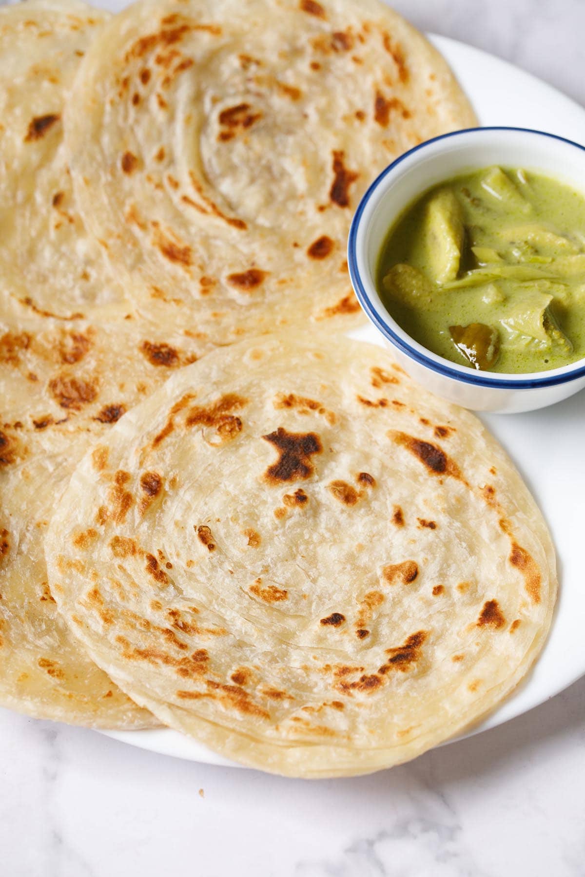 kerala parotta on white plate with a bowl of green curry at top right.