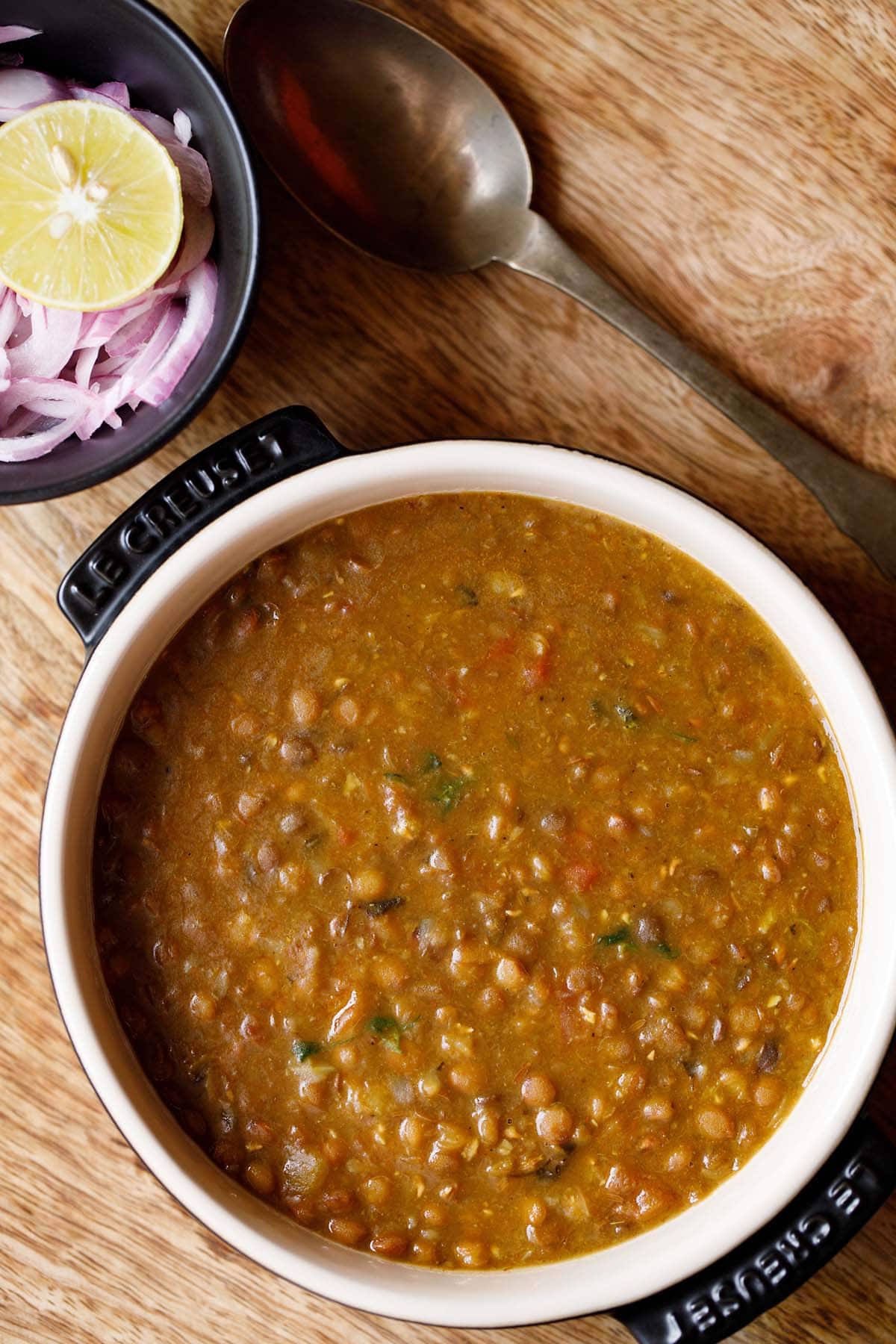 whole masoor dal served in a bowl with a bowl of onions and lemon kept in the top left and a spoon kept on the top right. 