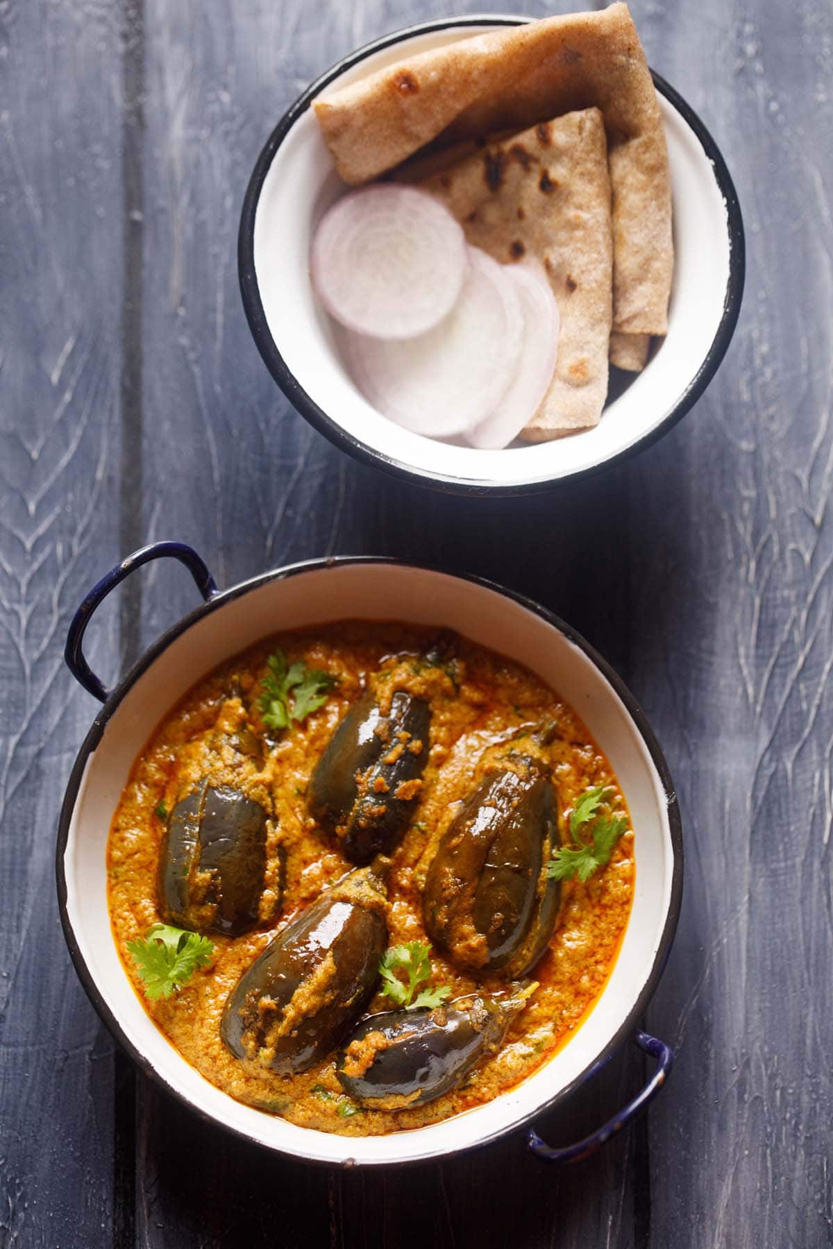 bharli vangi in a small enamelled white wok with a bowl of chapattis and onion slices kept in the background. 