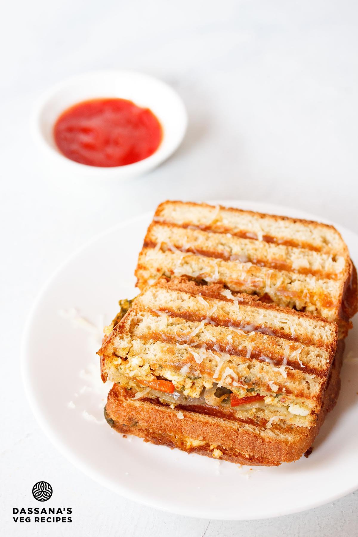 Paneer sandwich served on a white plate with a bowl of ketchup kept on the top left side.