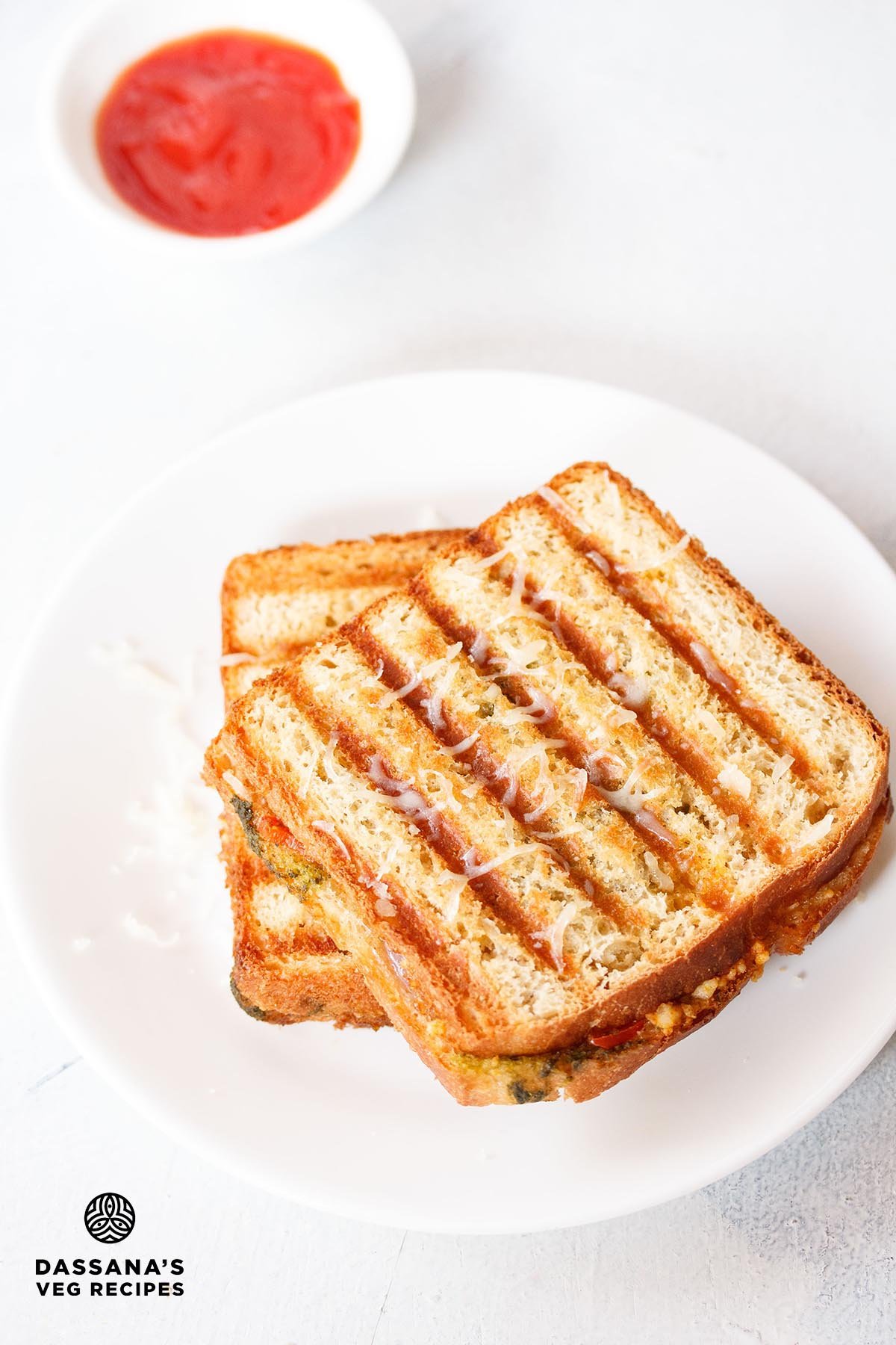 Grilled paneer sandwich on a white plate with a bowl of ketchup kept on the top left side.
