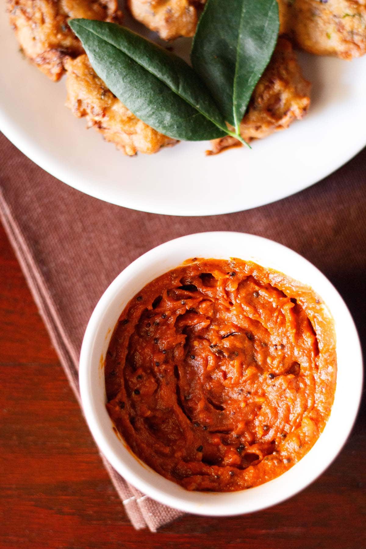 tomato chutney served in a small white bowl with a platter of pakoda kept on the top side.