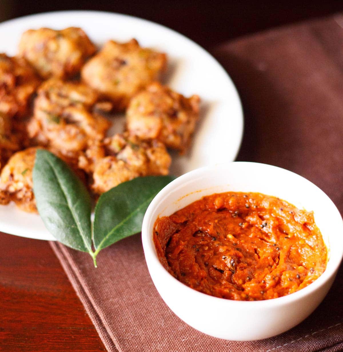 tomato chutney served in a small white bowl with a platter of pakoda kept on the top left side. 