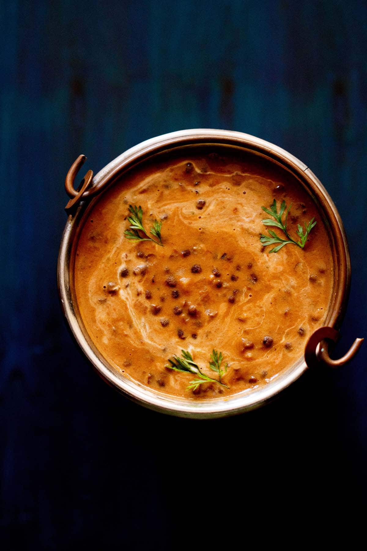 top shot of dal makhani garnished wit coriander sprigs served in a copper bucket on a dark blue board. 
