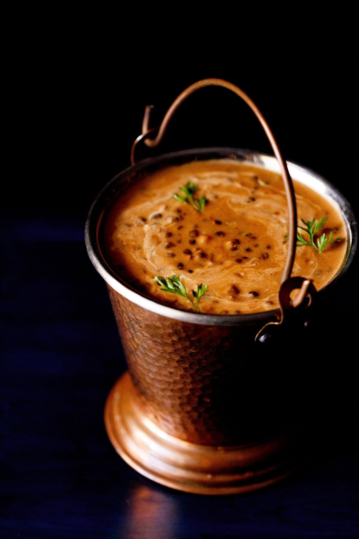 dal makhani garnished with coriander leaves and served in a fancy bucket with text layovers.