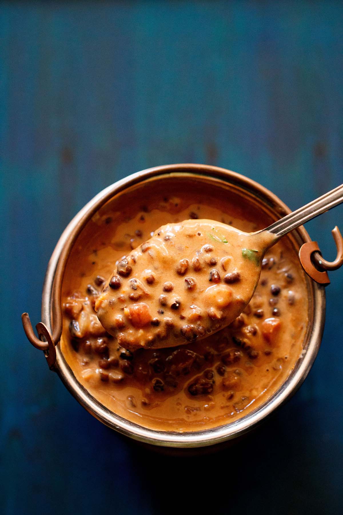 dal makhani served in a small copper bucket and taken in a spoon on a dark blue board. 