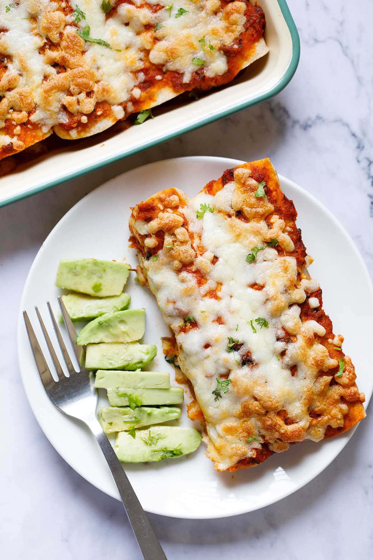 enchilada on a white plate with sliced avocado and fork by side.