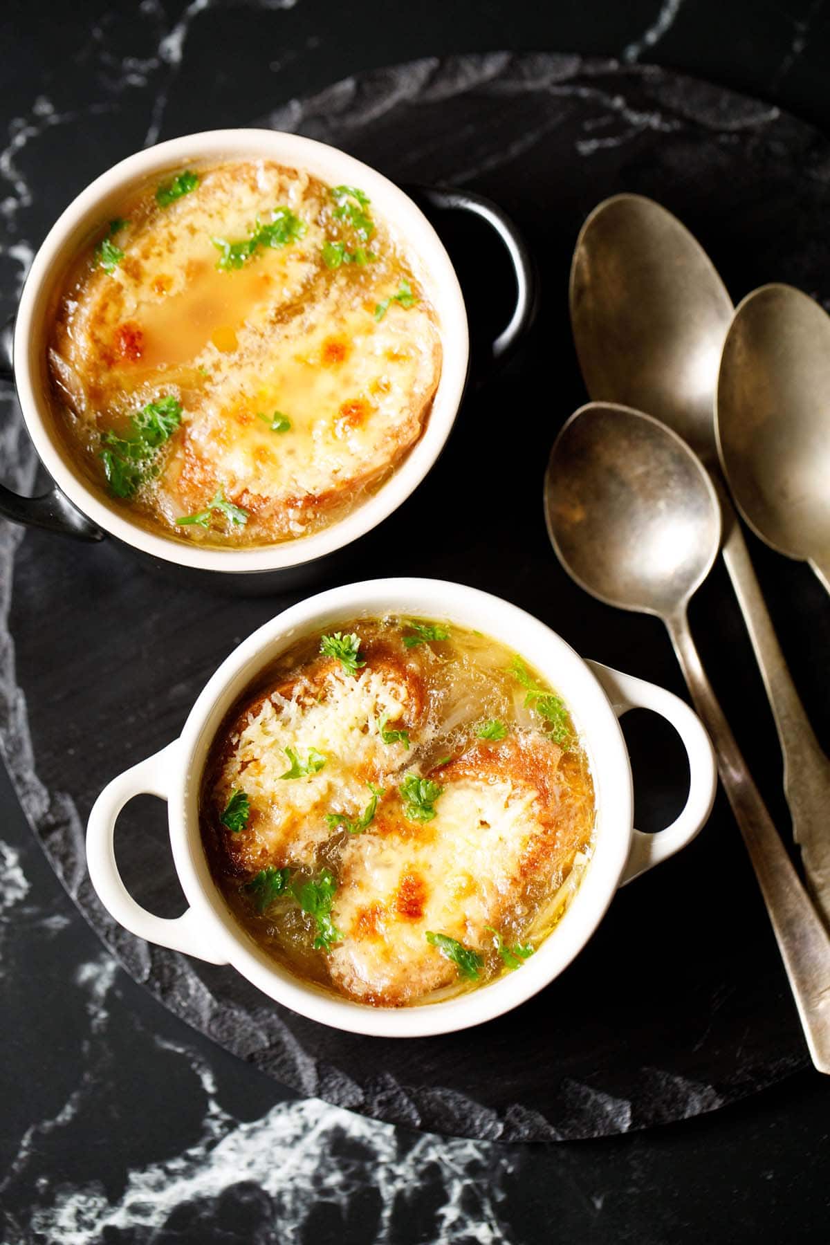 french onion soup in two cream colored soup bowls with spoons on a black slate tray. soup garnished with some parsley.