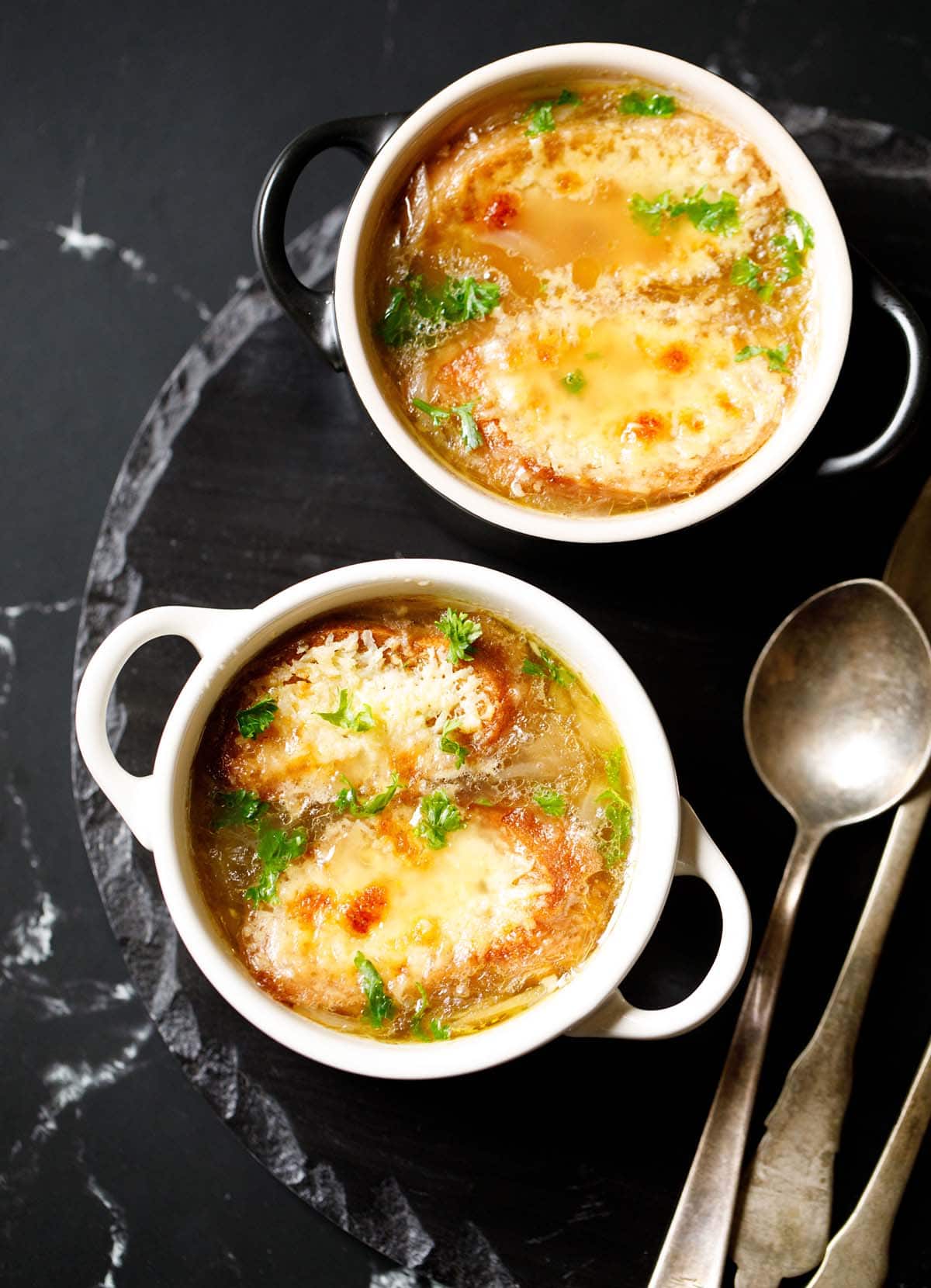 french onion soup in two cream colored soup bowls with spoons on a black slate tray. soup garnished with some parsley.