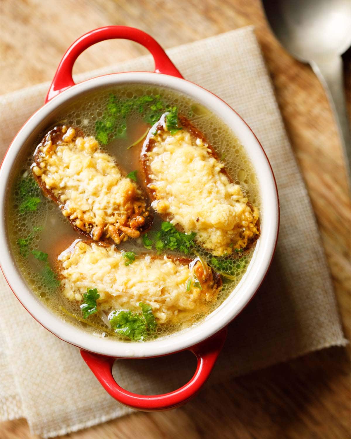 french onion soup in a cream soup bowl with red handles with toasted baguette slices with melted cheese on top.