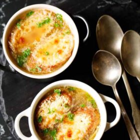 french onion soup in two cream colored soup bowls with spoons on a black slate tray. soup garnished with some parsley.