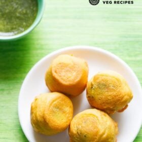aloo bonda served on a white plate with a small bowl of green chutney kept on the top side with text layovers.