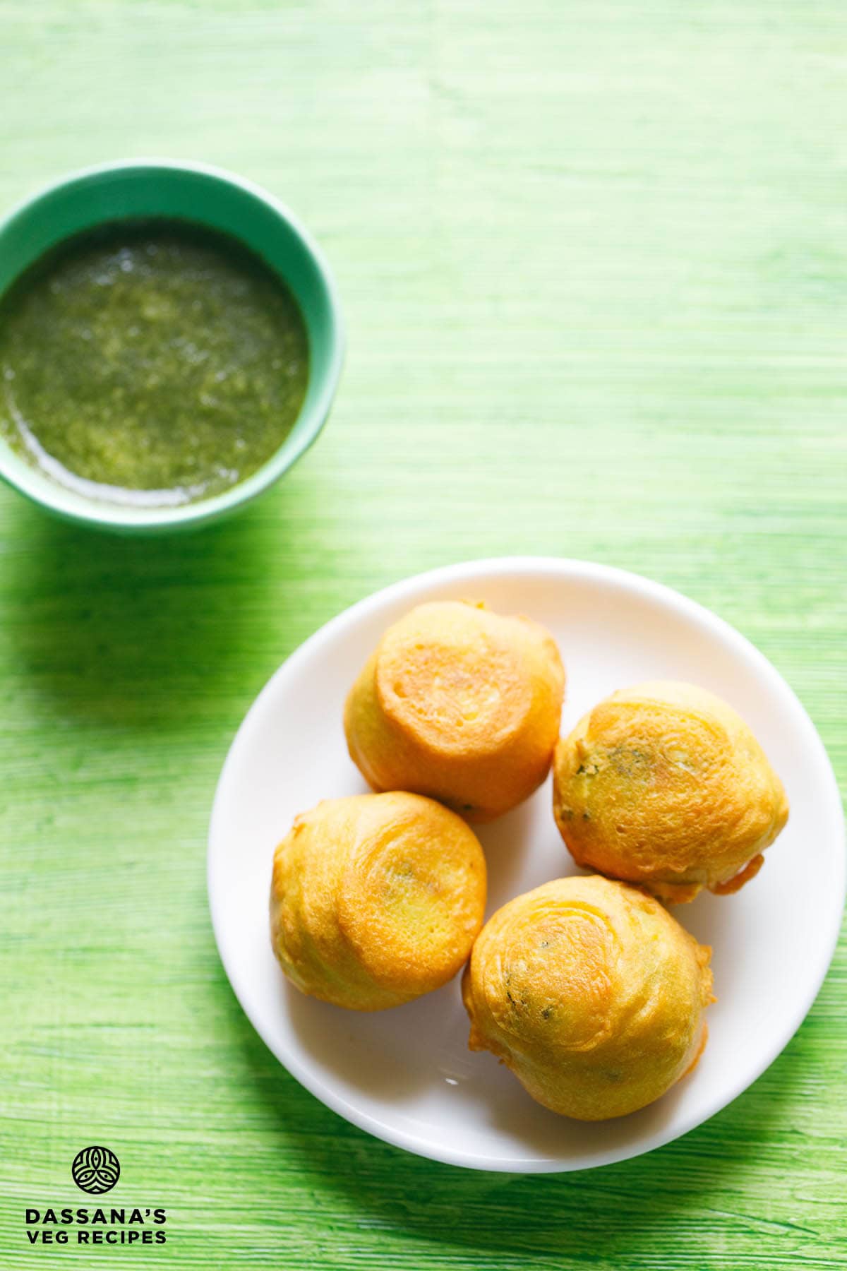 aloo bonda on a white plate with a small bowl of green chutney kept on the top side with text layovers.