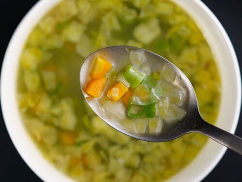 closeup image of clear soup with vegetables being seen in a steel spoon on top of white bowl with the soup on black table.