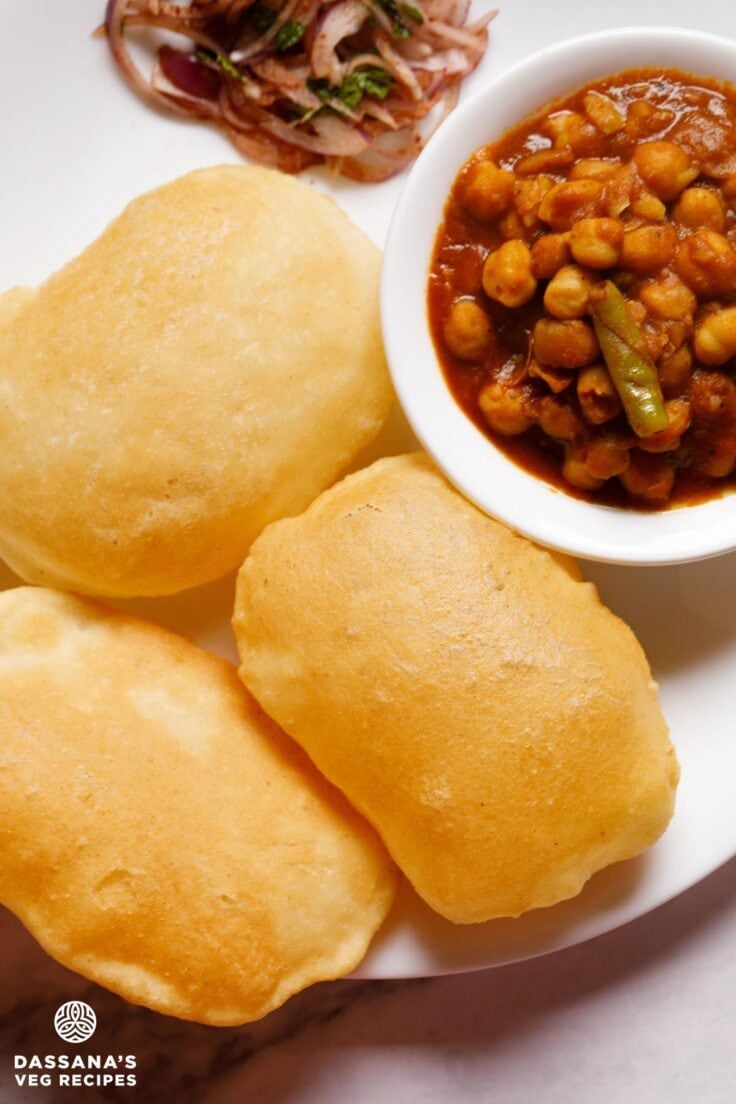 three bhatura with a side bowl of chana masala and some masala onions by the side on a white plate.