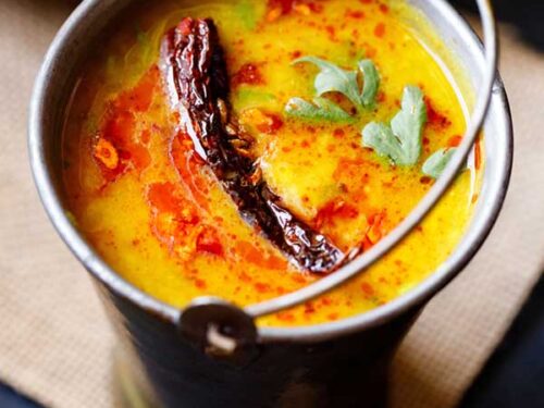 dal tadka garnished with cilantro and a topped with fried red chilli with some fried cumin and some red colored oil in a small brass bucket on a light brown jute mat.