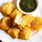 paneer pakora with one pakoda cut open showing the encased paneer inside, served on a white plate with green chutney in a small white bowl.