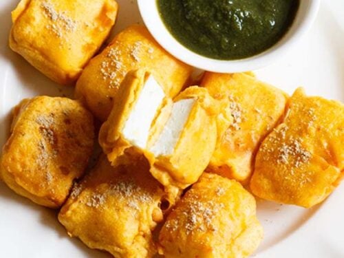 paneer pakora with one pakoda cut open showing the encased paneer inside, served on a white plate with green chutney in a small white bowl.
