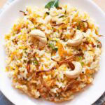 carrot rice garnished with cashews, fried onions and raisins on a white plate.