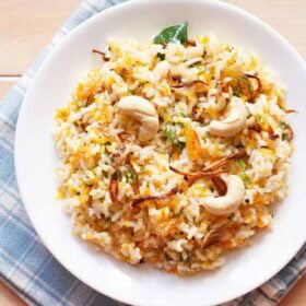 carrot rice garnished with cashews, fried onions and raisins served in a white plate on a checkered white and blue napkin with text layovers.