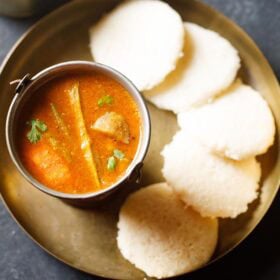 idli sambar combination with tiffin sambar in a bucket and five idli arranged in a semi-circle on a plate.