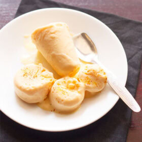 mango kulfi sliced and whole served on a white plate with a steel spoon.