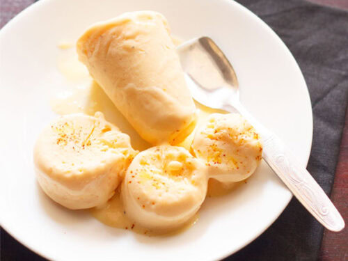mango kulfi sliced and whole served on a white plate with a steel spoon.