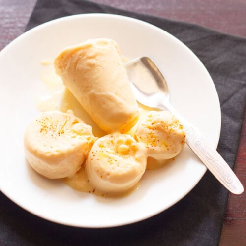 mango kulfi sliced and whole served on a white plate with a steel spoon.