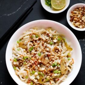 pad thai noodles in a white bowl garnished with crushed peanuts and spring onions with small bowls of crushed peanuts and chopped spring onions and lemon slices on top.