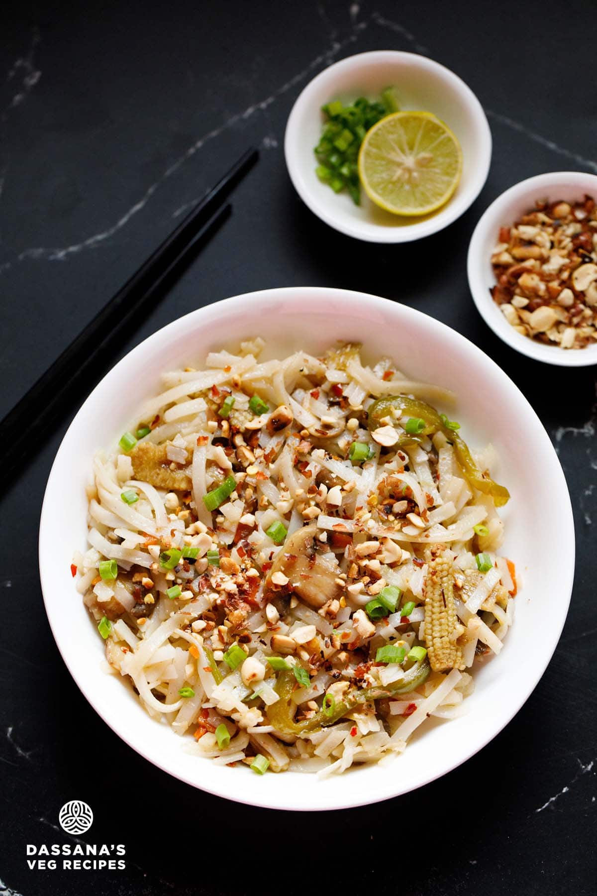 pad thai noodles in a white bowl garnished with crushed peanuts and spring onions with small bowls of crushed peanuts and chopped spring onions and lemon slices on top.