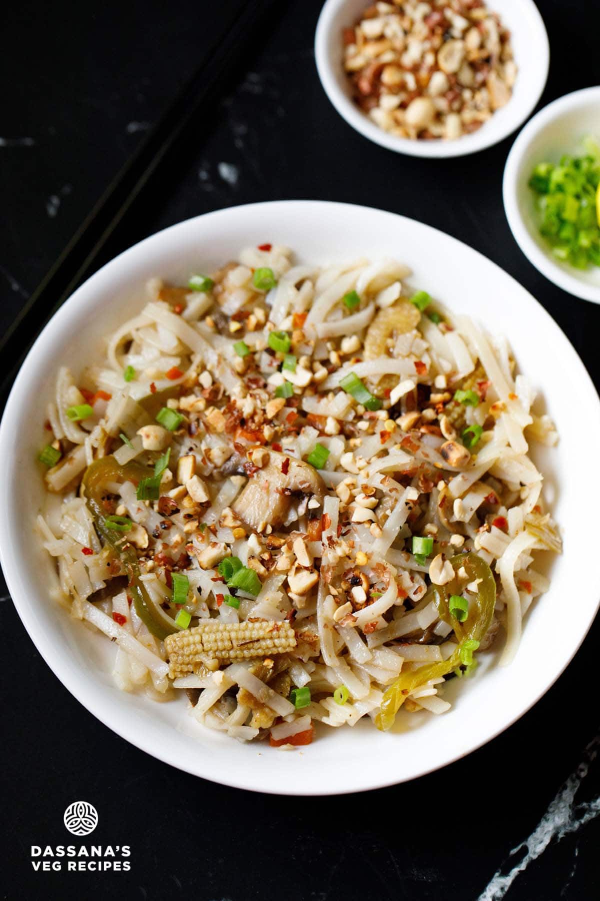 pad thai in a white bowl garnished with crushed peanuts and spring onions with small bowls of chopped spring onions & lemon slices and crushed peanuts on the top.