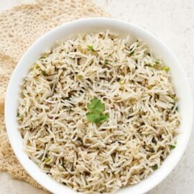 jeera rice in a white bowl with a coriander sprig in the center placed under a beige colored crocheted doily.
