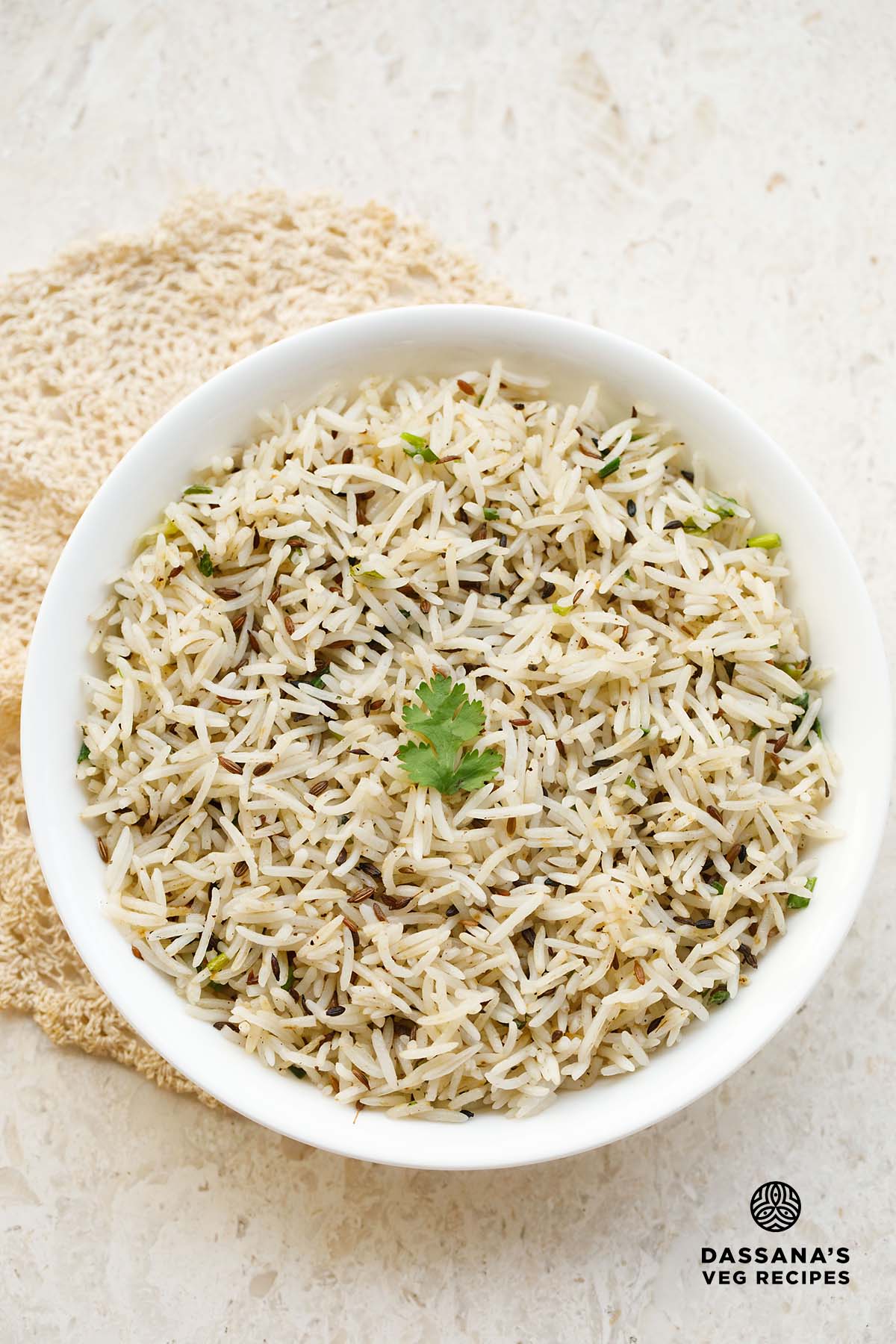 jeera rice in a white bowl with a coriander sprig in the center placed under a beige colored crocheted doily.