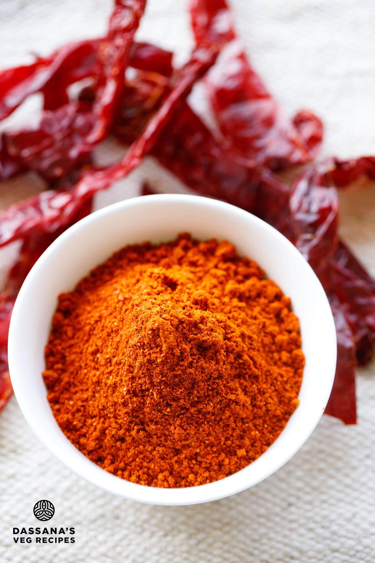 red chilli powder in a white bowl with dried red chillies on top and at the sides of the bowl on a jute cloth.